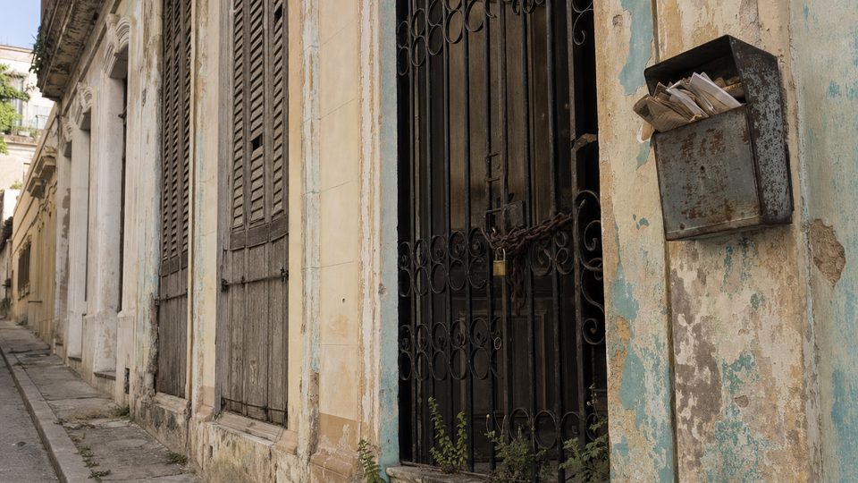 Casa habanera cerrada hace tiempo: contraventanas atrancadas, candado y cadena en la reja, hierbas en el umbral y el buzón desbordado